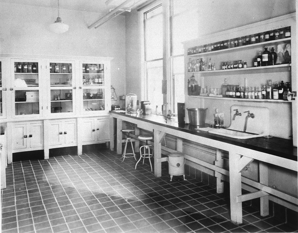 An empty bacteriology laboratory: a long bench with a sink and taps beneath a window, shelves of labeled bottles above it, glass-fronted cabinets on the left and a tiled floor.