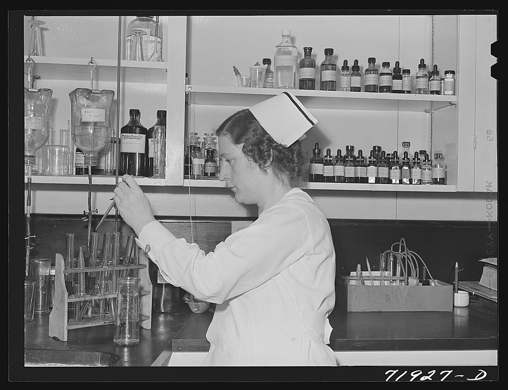 A nurse-technician in a white cap holds a test tube up to the light at a laboratory bench; racks of tubes and a shelf of reagent bottles surround her.