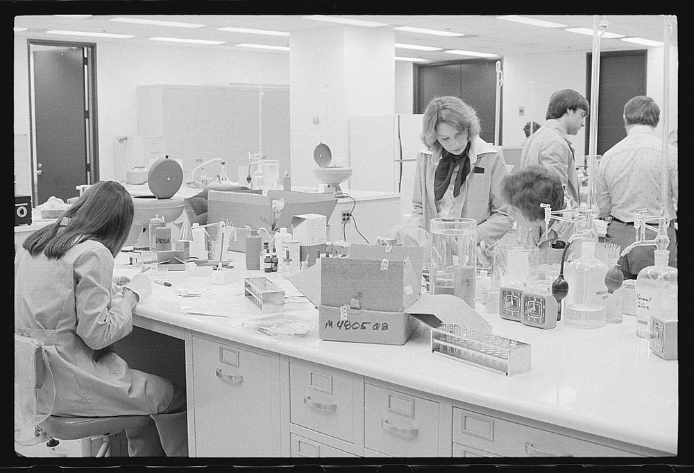 A busy 1970s laboratory: a woman with long hair sits at a bench in the foreground, colleagues work at benches behind her, and a cardboard box of samples and a rack of tubes sit on the counter.
