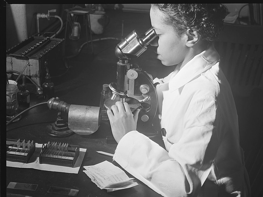 A young woman in a white coat looks into a monocular microscope at a dark bench; two wooden boxes of glass slides, a handwritten note, a pencil and small reagent bottles sit beside a microscope lamp, with a water bath and tubing on the counter behind. Archive photograph, 1942.