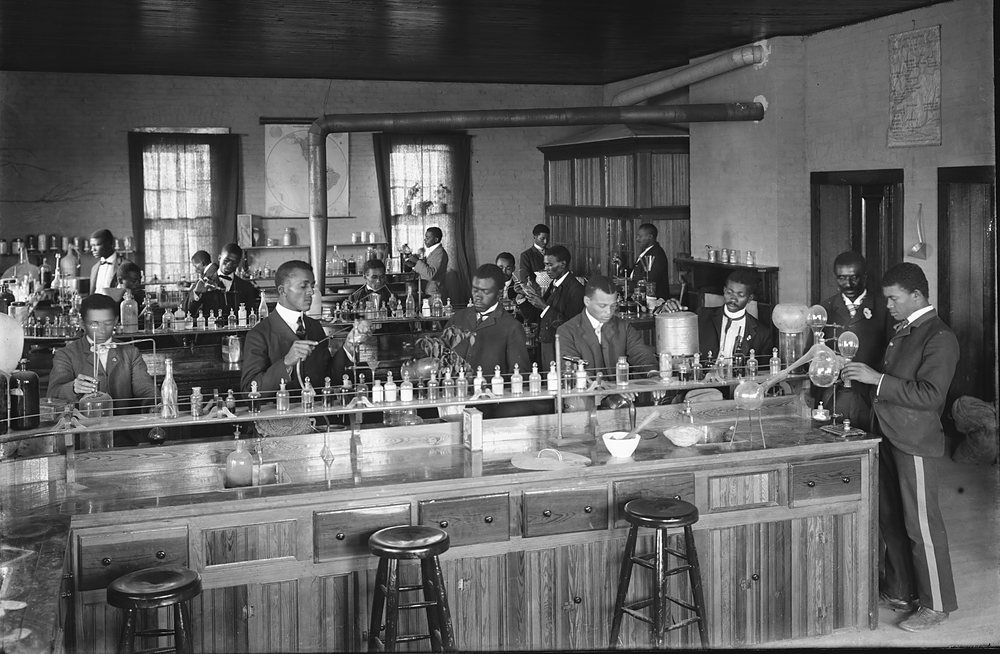 Young men in suits work at a long wooden chemistry bench at Tuskegee Institute, lined with rows of reagent bottles, flasks and a retort, beneath tall windows and ceiling pipes; wooden stools stand along the front of the bench. Archive photograph, 1902.