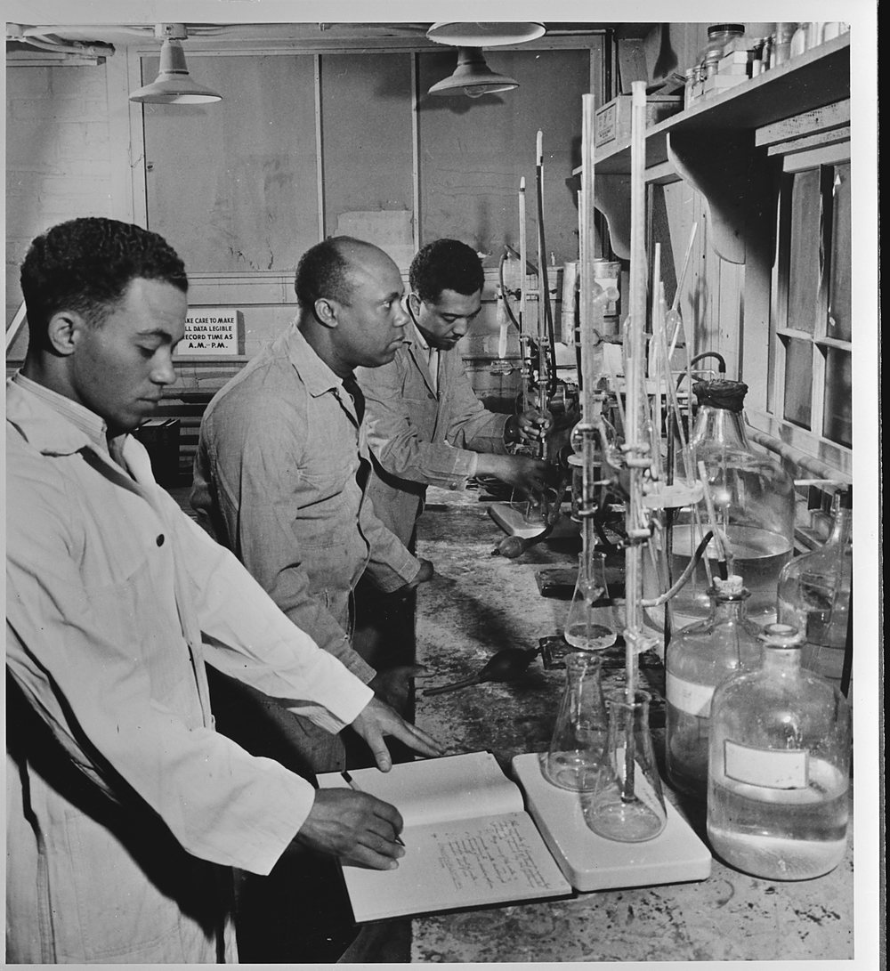 Three men stand in a line along a laboratory bench: in front a young man in a white coat writes in an open notebook, and behind him a bald man and a younger man work at tall stands of burettes, flasks and large glass bottles, under hanging lamps and wall shelves.