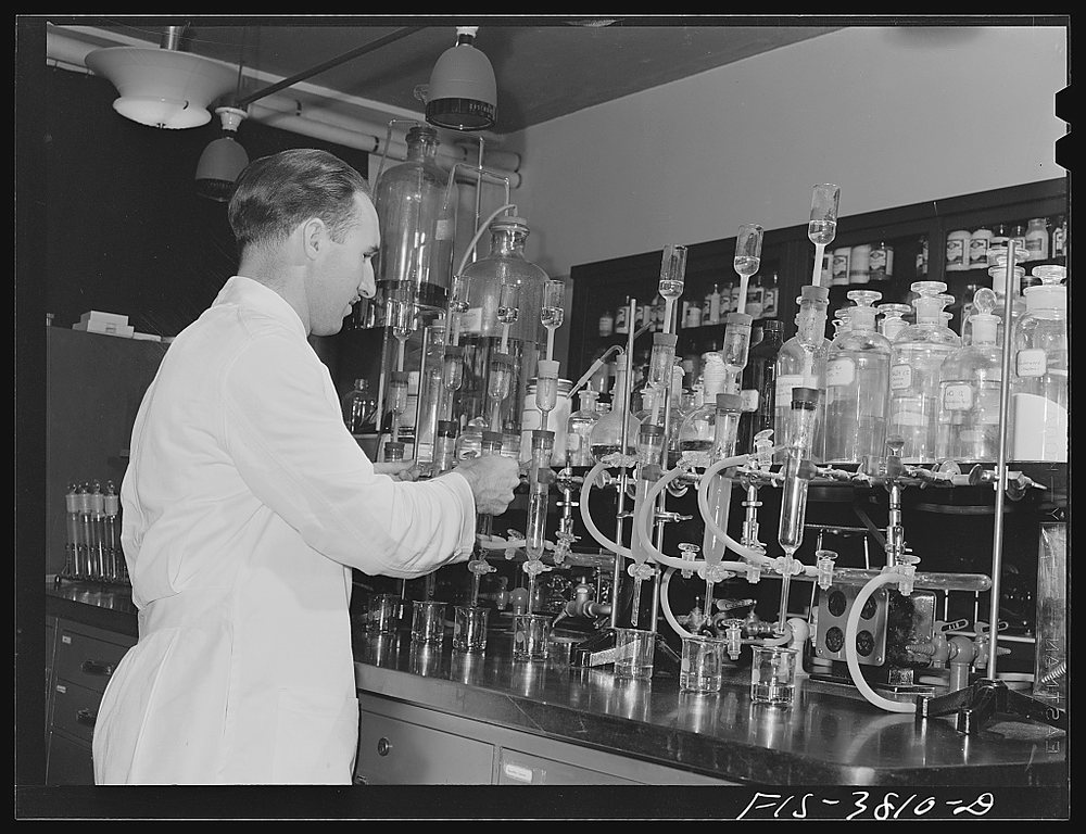 A man in a white coat works at a bench of tall glass apparatus, burettes and stoppered bottles beneath a shelf of reagent jars, under hanging pendant lamps.