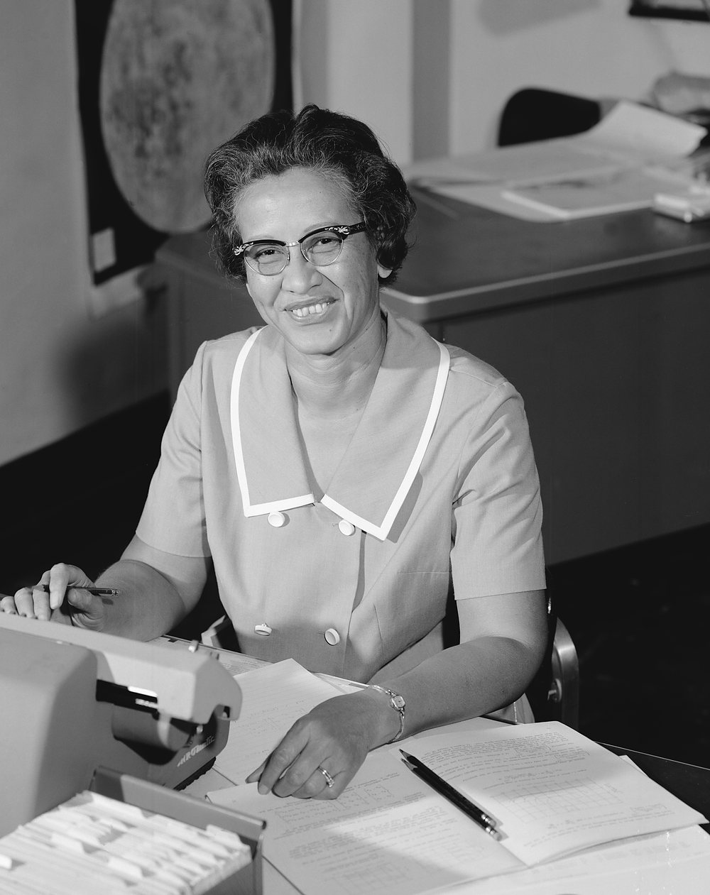 A woman in cat-eye glasses and a short-sleeved dress with a white-trimmed collar sits smiling at a desk, a pencil in her hand and handwritten worksheets in front of her; a mechanical calculating machine and a card file stand at the left, and a large photograph of the Moon hangs on the wall behind her. Archive photograph, 1966.