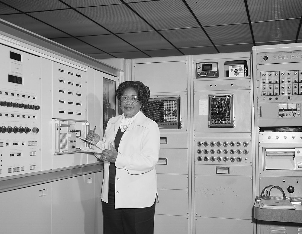 A woman in a white jacket stands smiling in front of a wall of instrument panels and racks, holding a sheaf of papers.