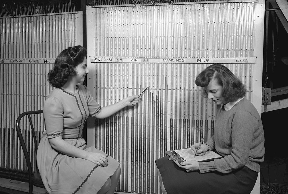 Two young women sit at a tall wall of manometer tubes in a wind-tunnel laboratory: one points a pencil at a liquid column while the other writes the reading on a data sheet on a clipboard in her lap. Archive photograph, 1943.