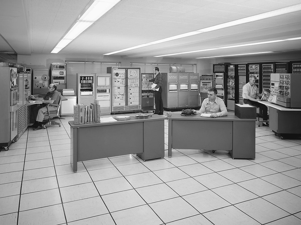 A 1970s data room: three men at gray desks and consoles among rows of tape drives and instrument racks under fluorescent panels, on a raised tile floor.