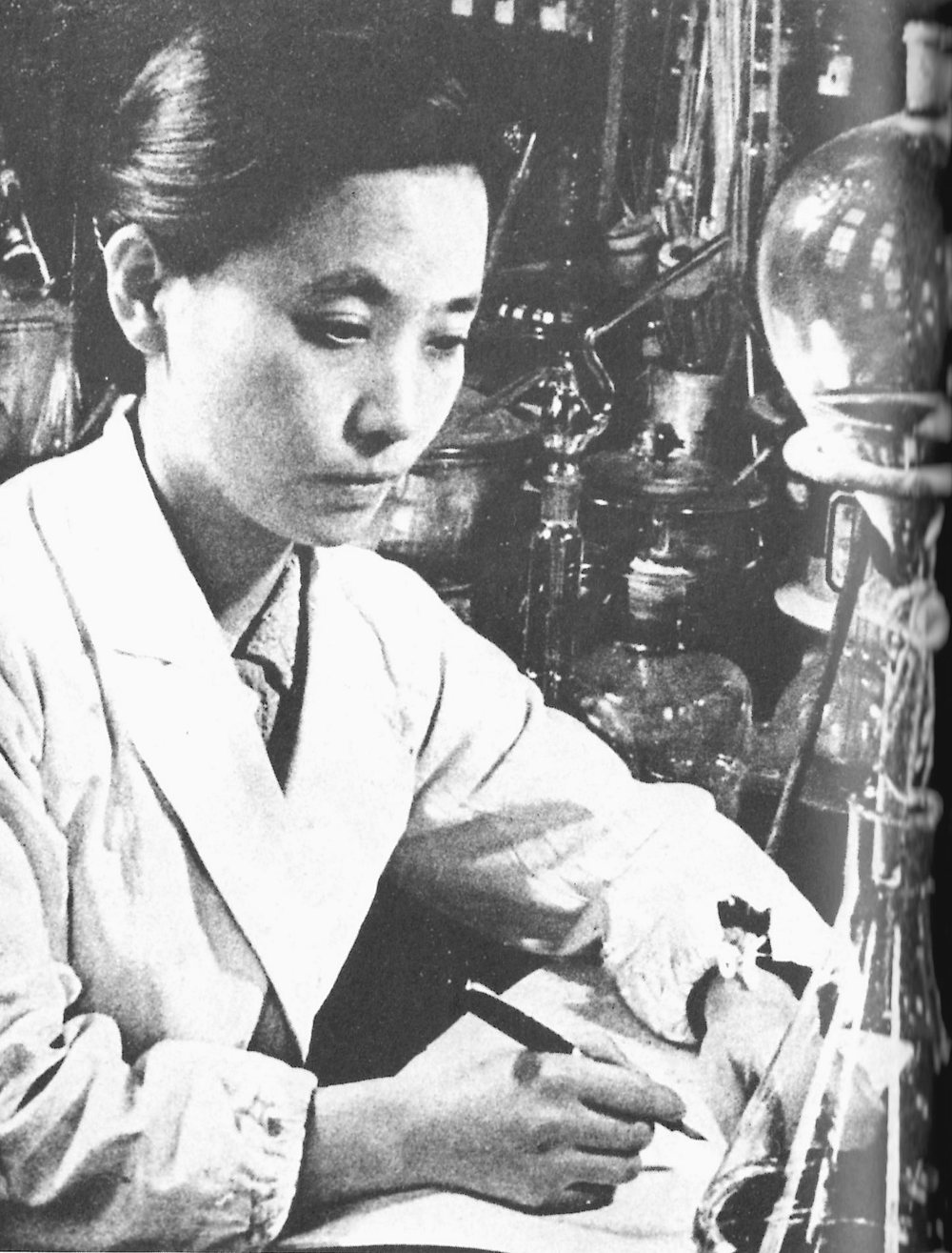 A woman scientist in a white coat writes in a notebook at her bench, surrounded by glass flasks and a tall condenser.
