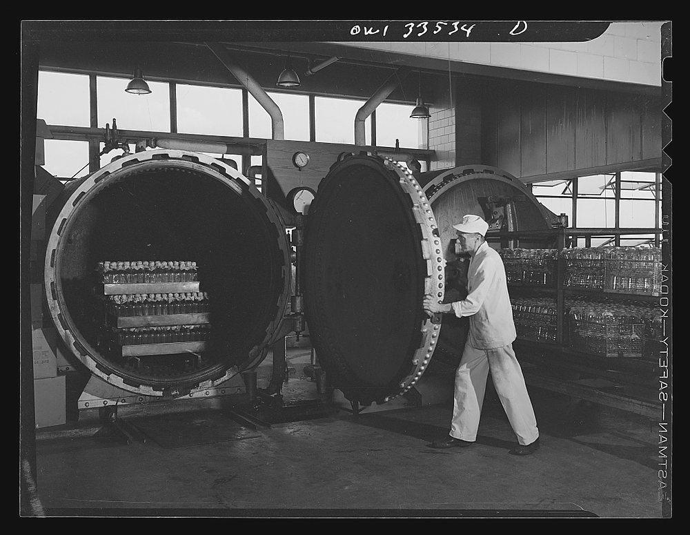 A worker in white swings open the round door of a large industrial autoclave; racks of glass containers wait inside and on a trolley beside him, under a row of pendant lamps.