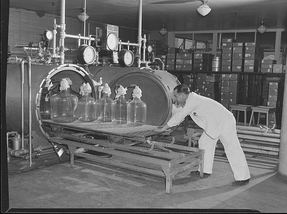 A man in white overalls pushes a wheeled rack of five large stoppered glass carboys into the open door of a horizontal steel autoclave fitted with pressure gauges and pipework; stacked crates fill the room behind him. Archive photograph, 1943.