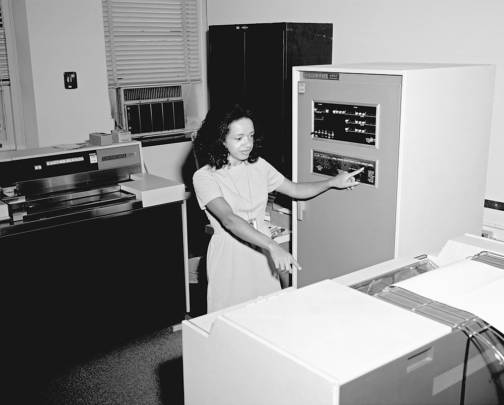 A woman in a short-sleeved dress stands in a computer room, pointing with one hand at the control panel of a tall computer cabinet and with the other toward a printer in the foreground; a second console stands at the left under a window with a blind and an air-conditioning unit. Archive photograph, 1973.