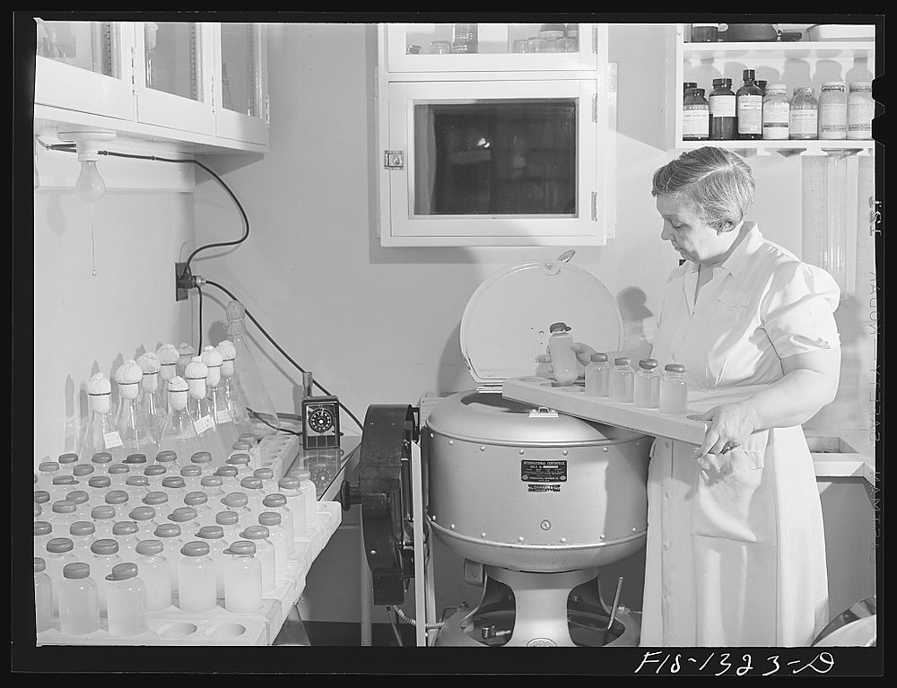 A woman in white lifts a tray of small bottles from an open centrifuge in a tiled laboratory; trays of stoppered bottles fill the bench to her left and a shelf of reagent jars hangs above.
