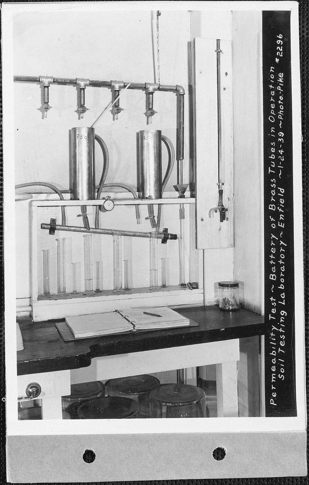 A battery of brass permeability tubes on a wall-mounted manifold above a row of graduated cylinders on a laboratory shelf; an open notebook and pencil lie on the bench below, and the print carries a handwritten caption dated 1-24-39.