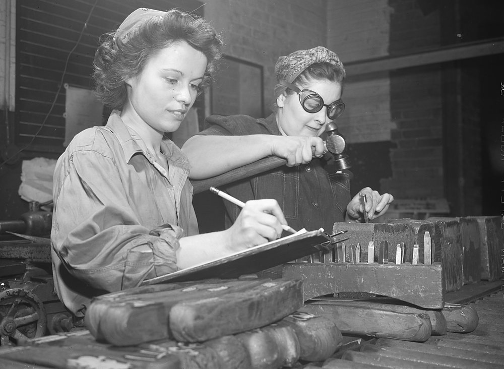 A young woman in a work shirt and headscarf writes on a clipboard at a bench of steel test samples while an older woman in dark goggles stamps identification numbers onto a rack of small test bars with a hammer and punch; heavy billet samples lie in the foreground.