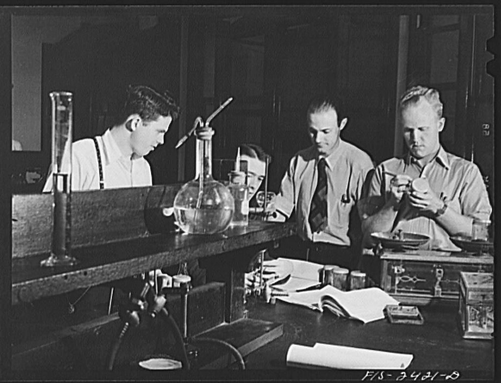 Four young men at a soils laboratory bench: one leans over a burette and flask, two confer over an open notebook and papers, a fourth weighs a sample on a balance beside a wooden case.