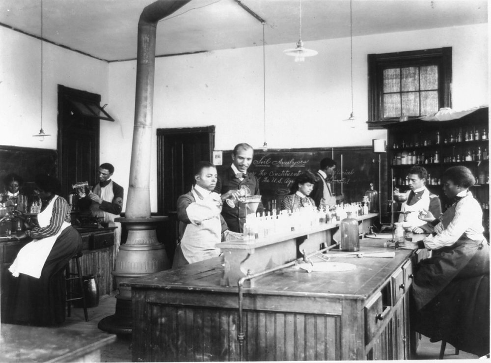 Students and an instructor at wooden benches in a soil-analysis teaching laboratory at Tuskegee Institute: at the center bench a man in a suit pours from a beaker while a young man in an apron watches, women in long skirts and white aprons work at the benches on either side, and a blackboard headed Soil Analyses hangs behind them beside shelves of reagent bottles. Archive photograph, 1902.