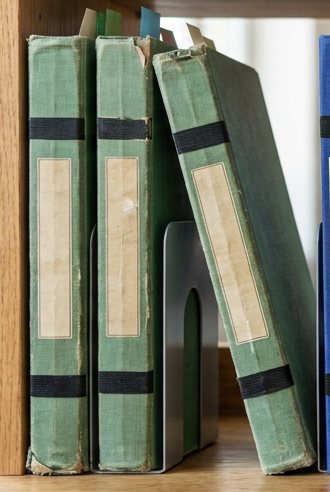 Digitally rendered: three faded green hardbound notebooks with blank cream labels stand on a wooden shelf, the third leaning on the second against a metal bookend.