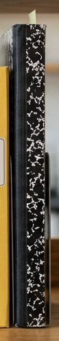 Digitally rendered: the spine of a black marbled cloth composition book on a wooden shelf.