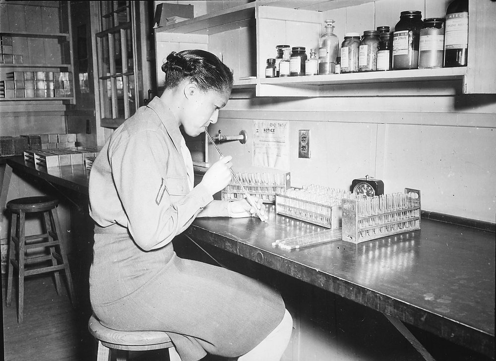 A woman in an Army uniform sits on a stool at a long black bench, pipetting by mouth into a rack of test tubes; more tube racks and a small clock stand on the bench and a shelf of labeled reagent jars hangs above.
