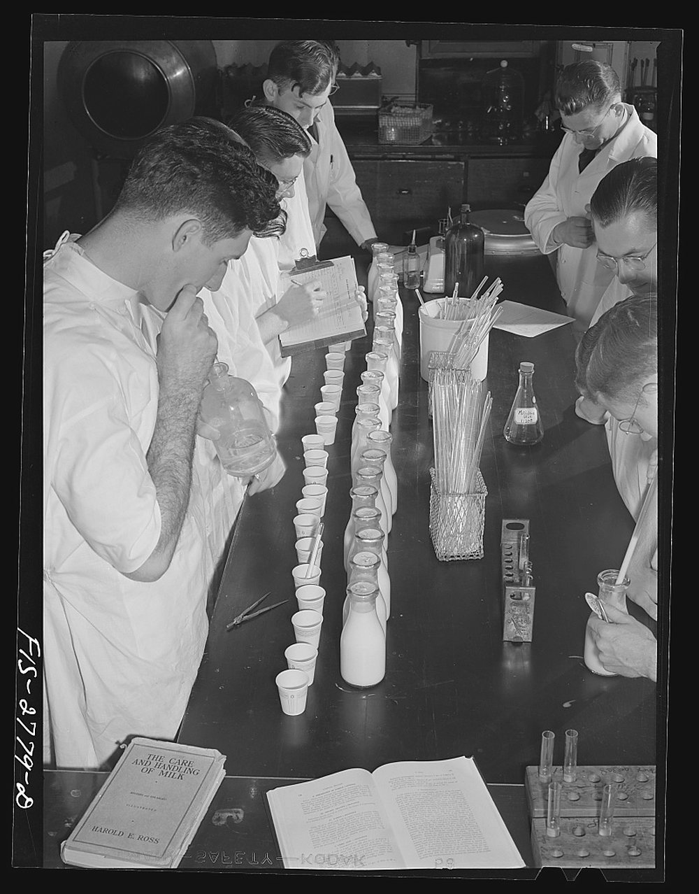 Five students in white coats crowd a dark bench covered with rows of small paper cups and milk bottles; one writes on a pad, another reads, and a textbook lies open at the front.