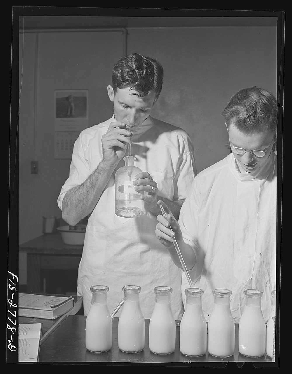Two senior veterinary students in white coats test milk samples at a bench: one draws from a glass bottle with a pipette, the other holds a long pipette over a row of milk bottles; a calendar hangs on the wall behind them.